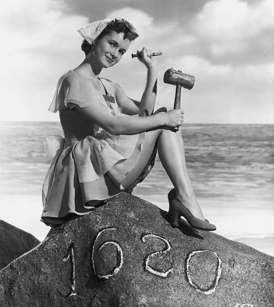 Debbie Reynolds, dressed as a pilgrim, sits on top of a replica of Plymouth Rock holding a mallet and a chisel.