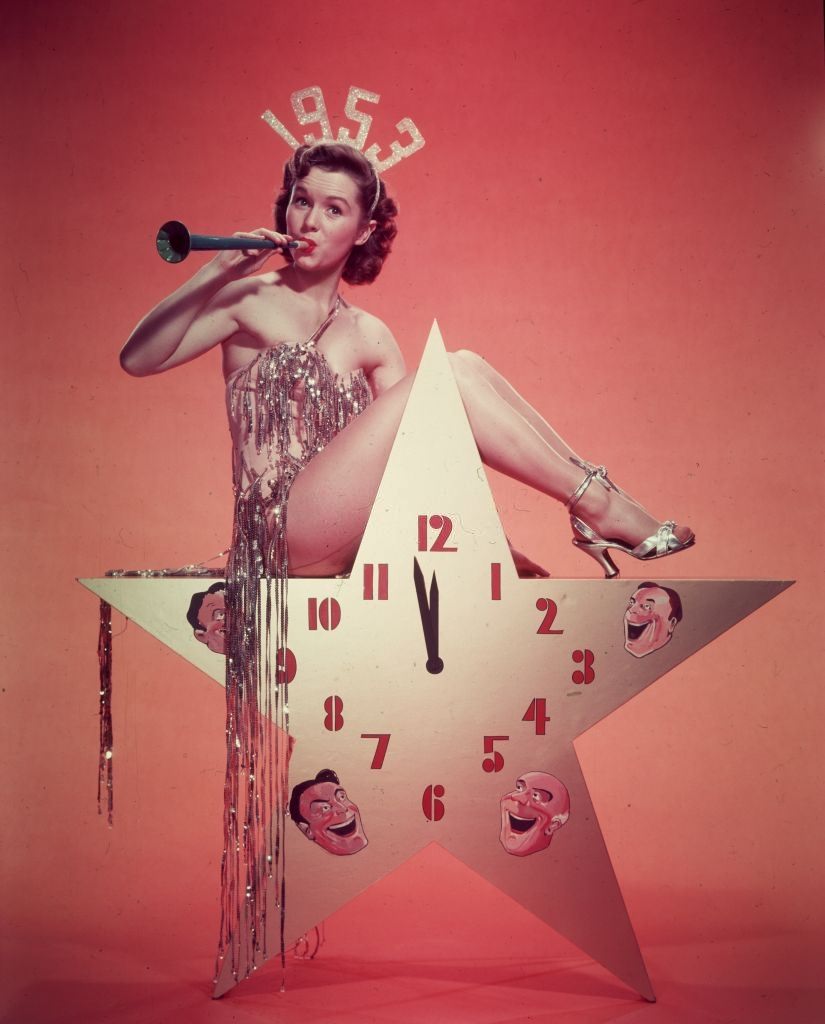 Debbie Reynolds, wearing a 1953 tiara and a costume with tinsel, blows a horn while sitting atop a star-shaped clock in a promotional portrait for New Year's Eve.