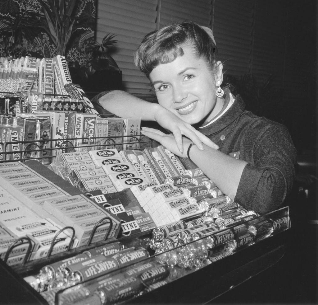 Debbie Reynolds smiles while leaning upon a counter filled with a display of various candies, 1952.