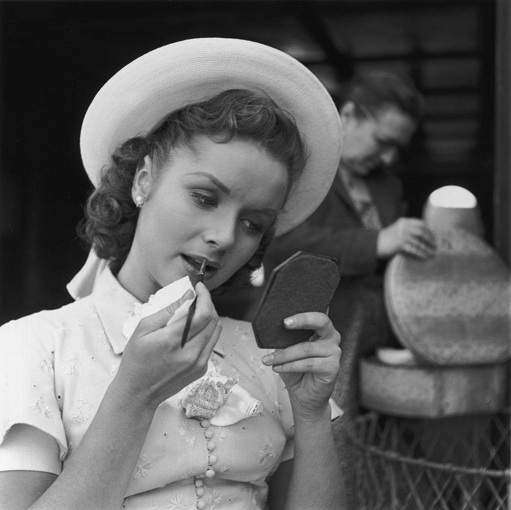 Debbie Reynolds, wearing a straw hat, applying lipstick behind the scenes of director Don Weis's film, 'I Love Melvin', 1952.