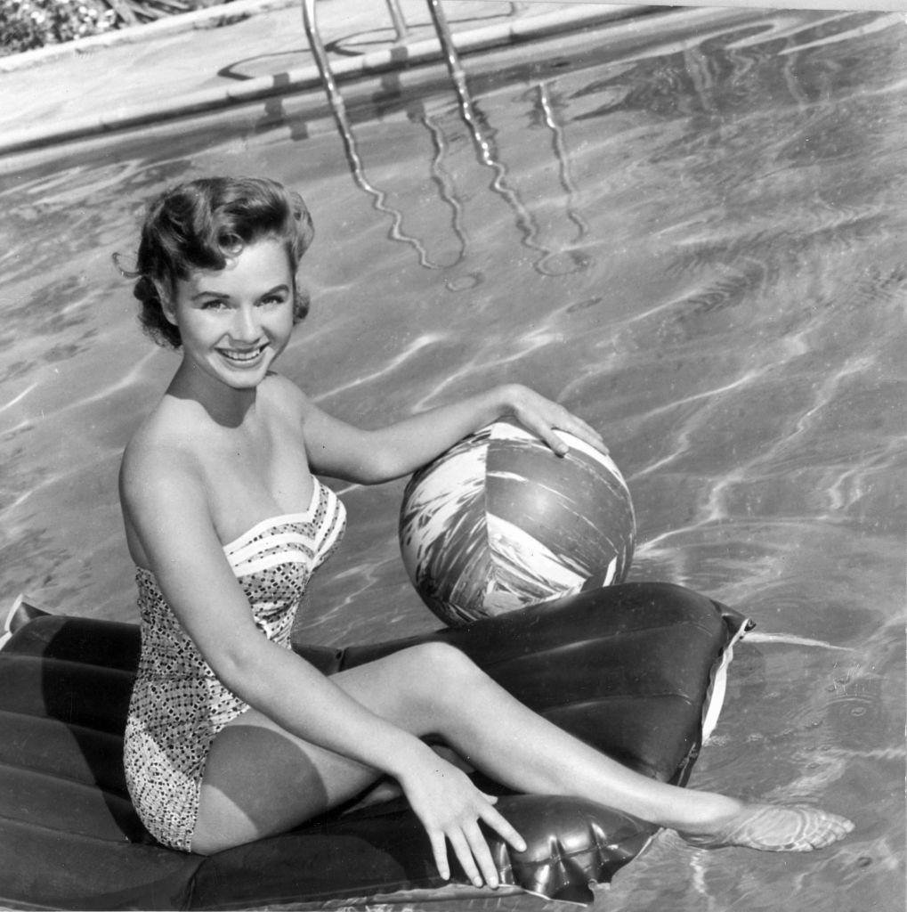 Debbie Reynolds, wearing a strapless one-piece swimsuit, holding a beach ball while sitting on an air mattress in a swimming pool, 1955.