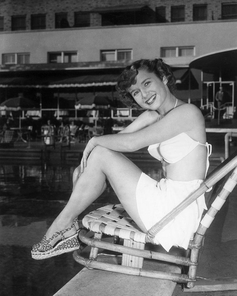 Debbie Reynolds, smiling and sitting in a beach chair at the edge of a swimming pool, 1955.
