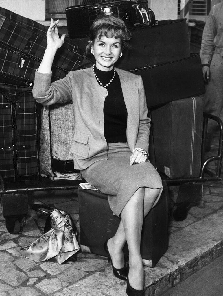 Debbie Reynolds sits on a piece of luggage, waving and smiling, 1955.