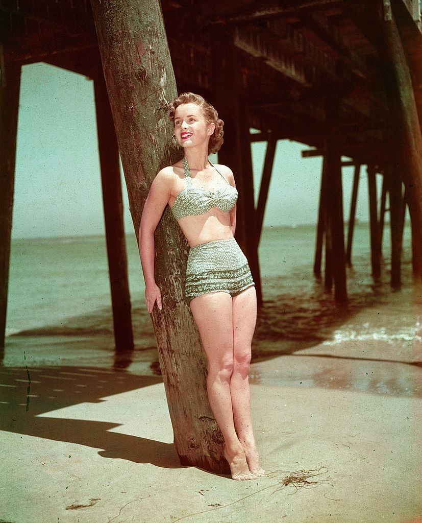 Debbie Reynolds wearing a two-piece swimsuit while leaning against a wooden pier post on a beach, 1950s.