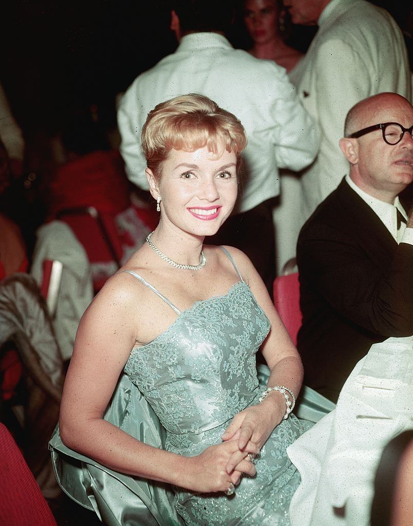 Debbie Reynolds, wearing an evening dress, smiles while sitting in the crowd at a formal event, 1950s.