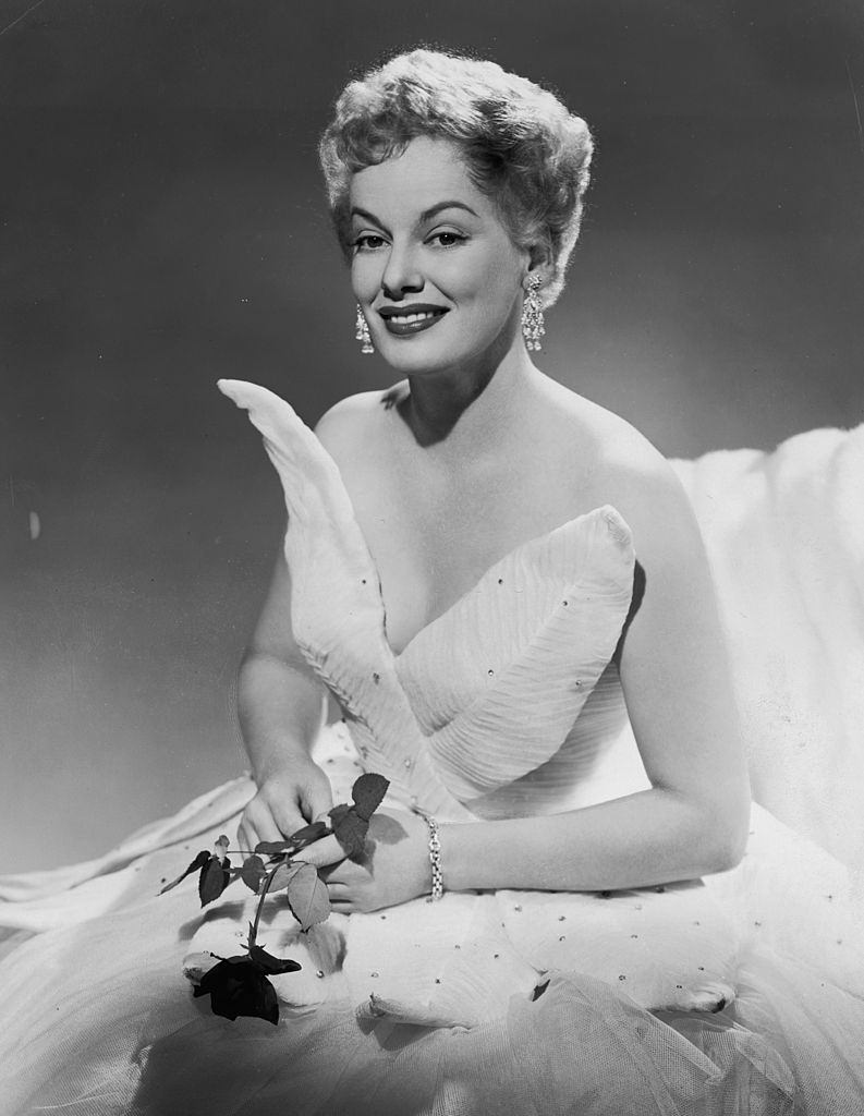 Faye Emerson wearing a leaf-patterned white gown, and holding a rose, 1948.