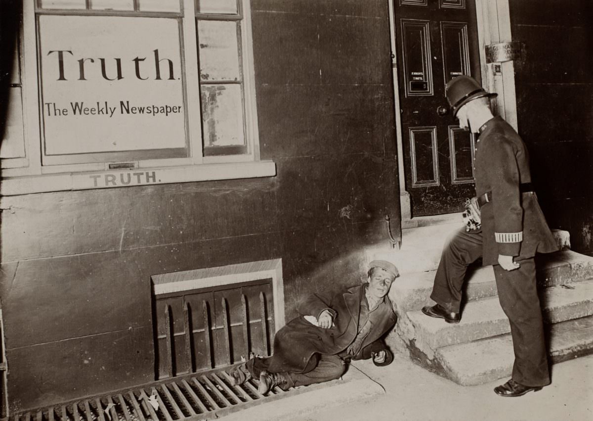 “A policeman shines a flashlight onto a young man sleeping on the sidewalk against a building.