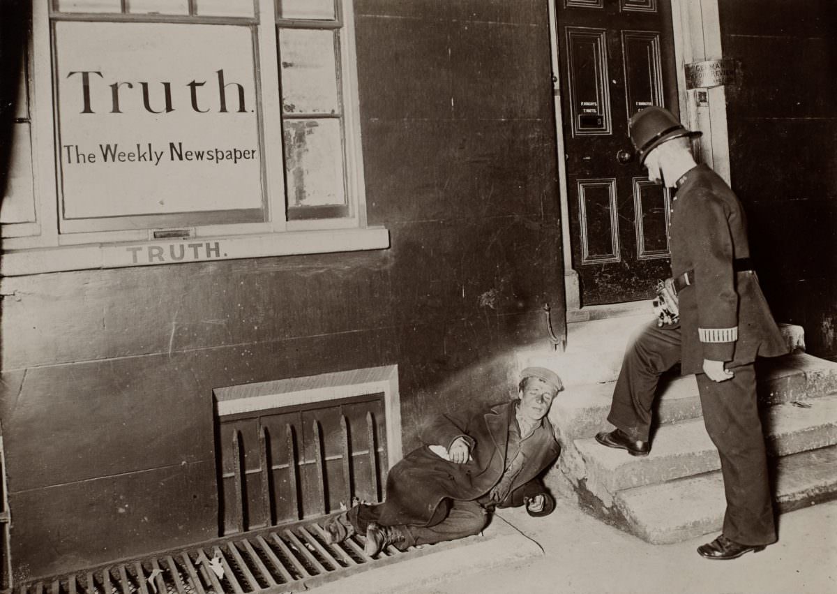 “A policeman shines a flashlight onto a young man sleeping on the sidewalk against a building.
