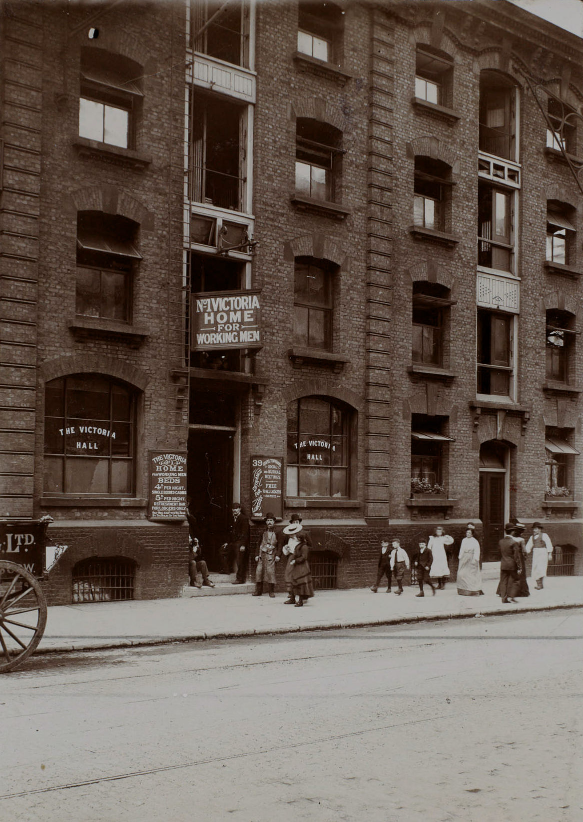 A group of men stand out on the stoop of a four-story brick building, while a few women and children walk by on the sidewalk.