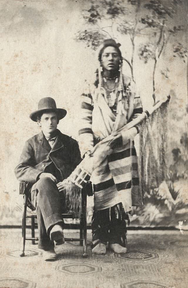 Young Native American man with a rifle standing next to a seated Caucasian man wearing a hat