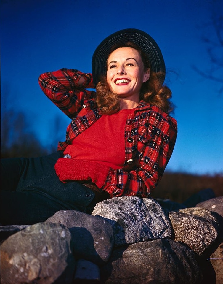 Paulette Goddard poses atop rocks with her hand on her hat.
