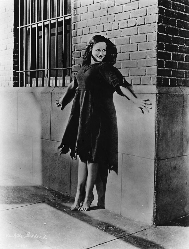 Paulette Goddard standing barefoot and on tiptoe in front of a barred window, 1936.