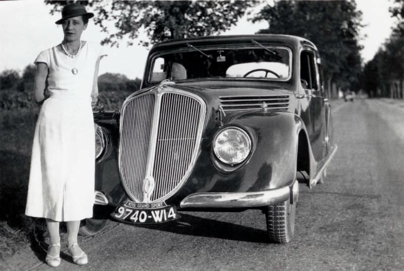 An elegant lady in a white dress posing with a Renault Viva Grand Sport on a tree-lined avenue in summertime, circa 1936