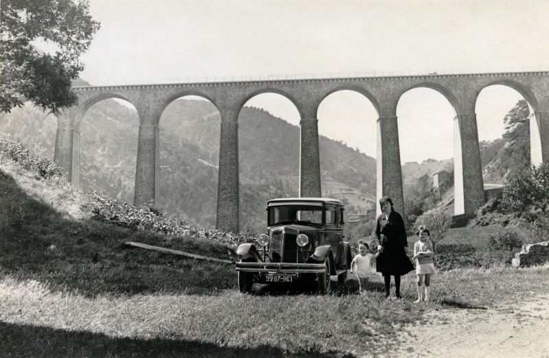 A mother and her children posing with a Renault Monasix in a valley dominated by a distinctive arched bridge, possibly in the South of France.