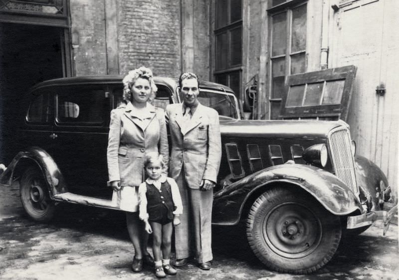 A family of three posing with a somewhat battered Renault Vivastella in an urban backyard, 1948