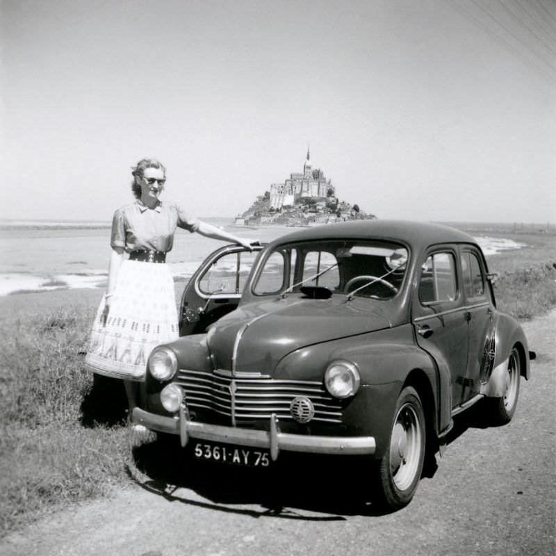 A blonde lady posing with a Renault 4 CV in front of the impressive silhouette of a famous landmark in Normandy.