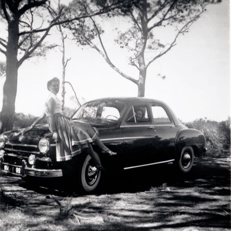 A lady wearing a 3/4 length skirt and a white sleeveless top casually posing on the fender of a Renault Frégate in summertime.