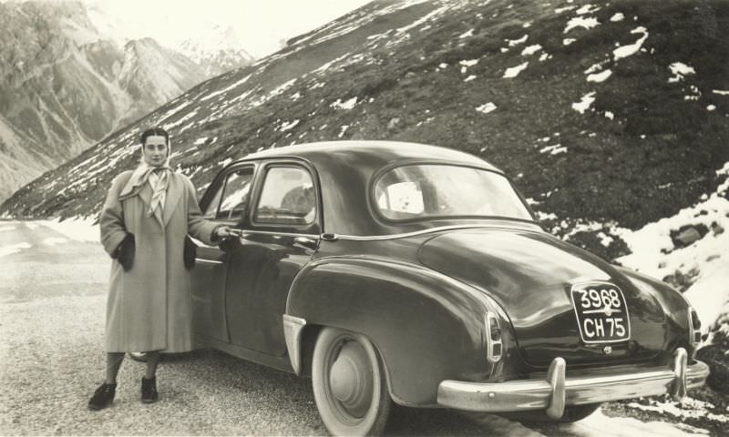 A lady wearing a silk headscarf and a woollen coat posing with a Renault Frégate on a mountain road.