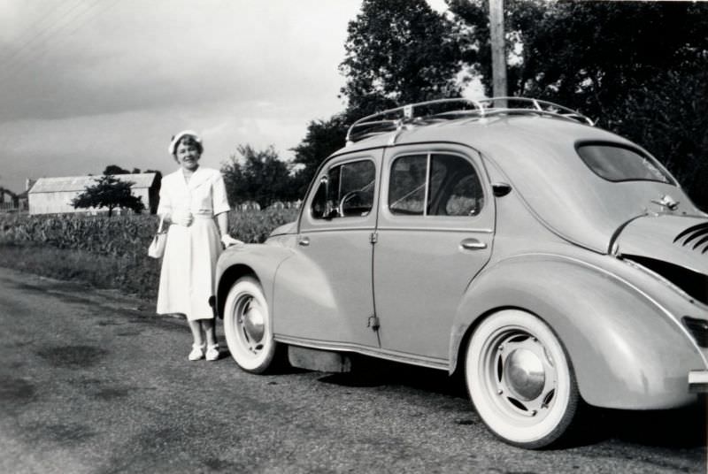 A stylish lady in a white dress with matching beret, handbag, and shoes posing with a Renault 4 CV in the countryside, 1954