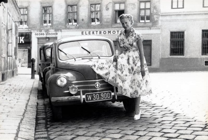 A blonde lady in a floral dress posing with a Renault 4 CV in a cobbled street in summertime.