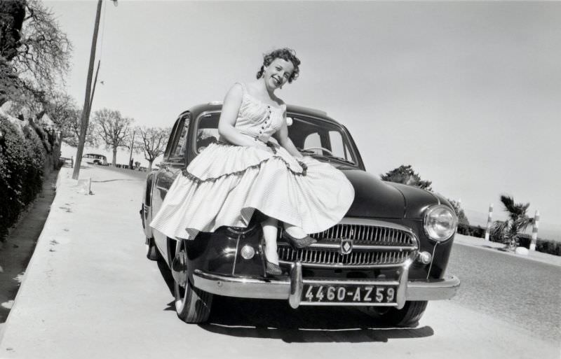 A cheerful lady posing on the fender of a Renault Frégate on a coastal road in mid-day sunshine.