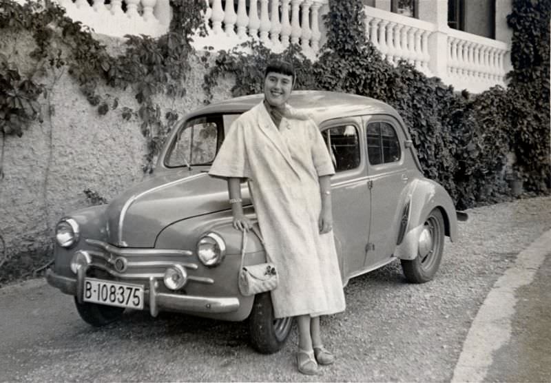 A cheerful lady posing with a Renault 4 CV in front of an ivy-clad balcony.
