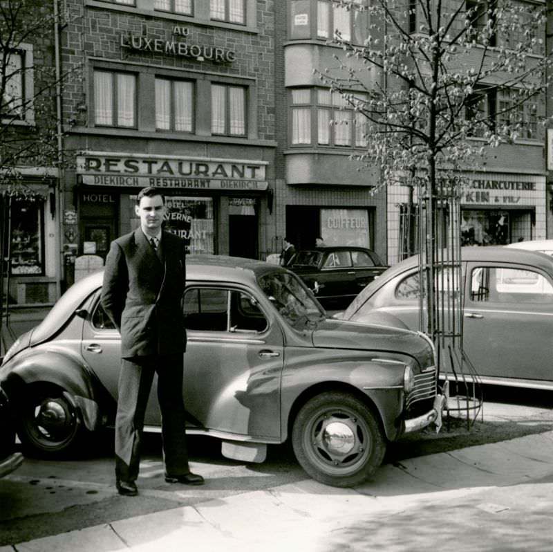 A dapper fellow in a double-breasted suit posing with a Renault 4 CV in a small town high street, Diekirch, Luxembourg, 1955