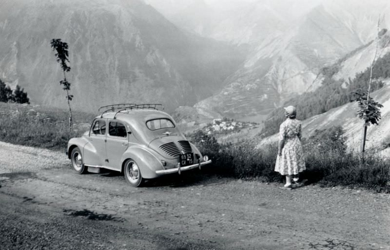 A lady in a floral dress posing with a Renault 4 CV on an Alpine road in summertime.