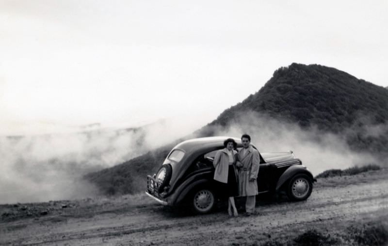 An adventurous couple posing with a Renault Juvaquatre on a gravelled mountain road on an overcast day, 1955