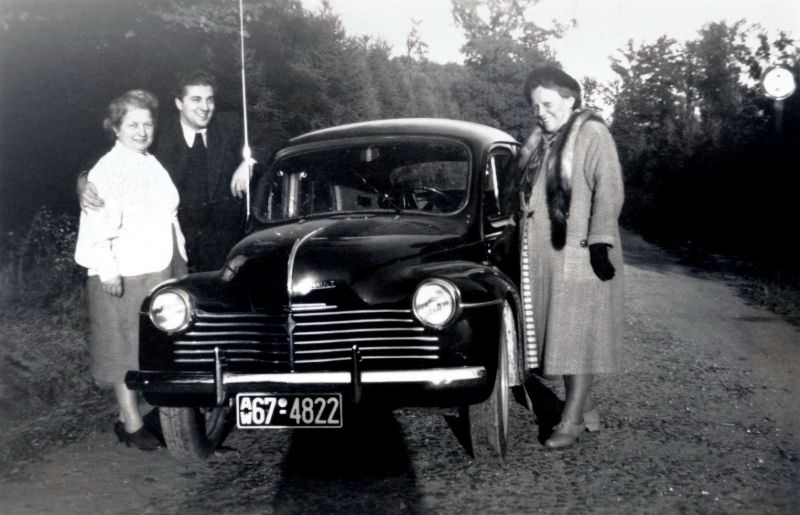 Three members of a middle-class family posing with a Renault 4 CV on a gravel road.