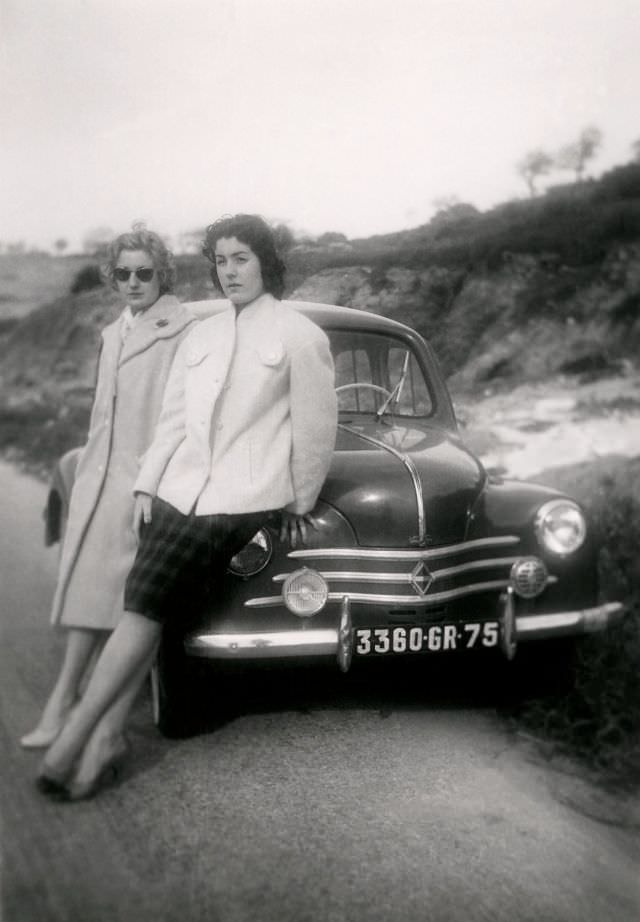 Two fashionable ladies posing with a Renault 4 CV on the side of a road in the countryside.