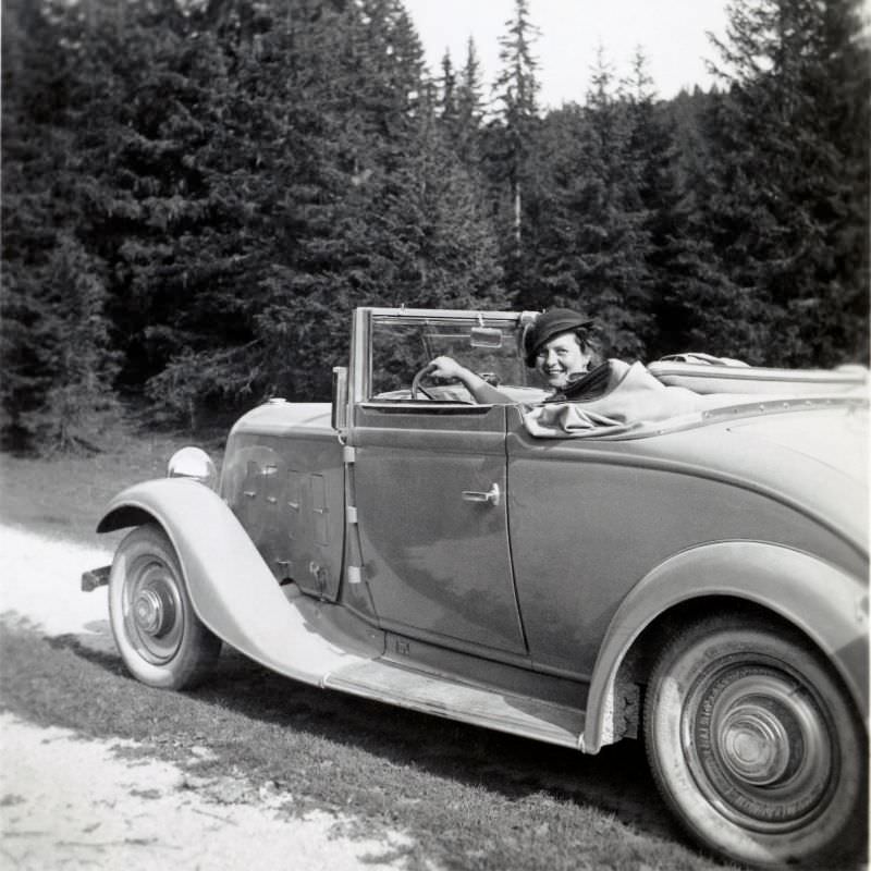 A cheerful lady posing in the driver's seat of a Renault Monaquatre Cabriolet-Spider convertible on a dirt road in the countryside, 1935