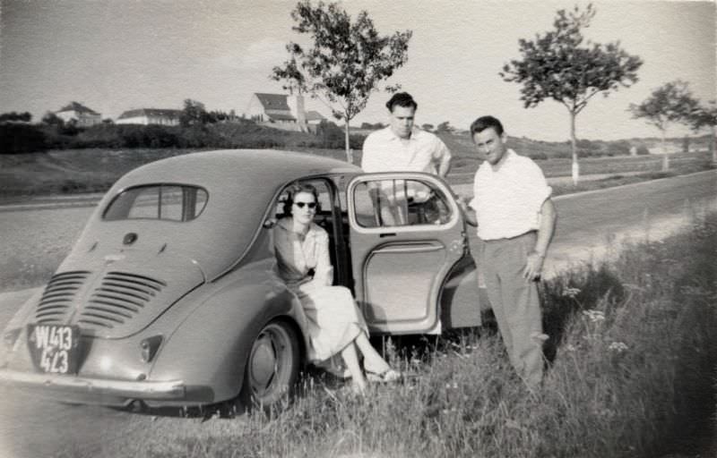 A company of three posing with a Renault 4 CV on a country road in summertime. The car is registered in the city of Vienna, 1957
