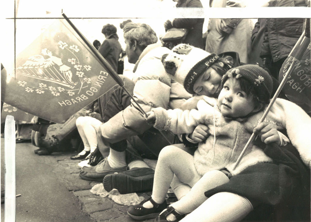 Snuggled together at their curbside seats, parade celebrants Mary Kathleen Collins, 2, and her sister Noreen, 9, wave Irish flags. March 1979.