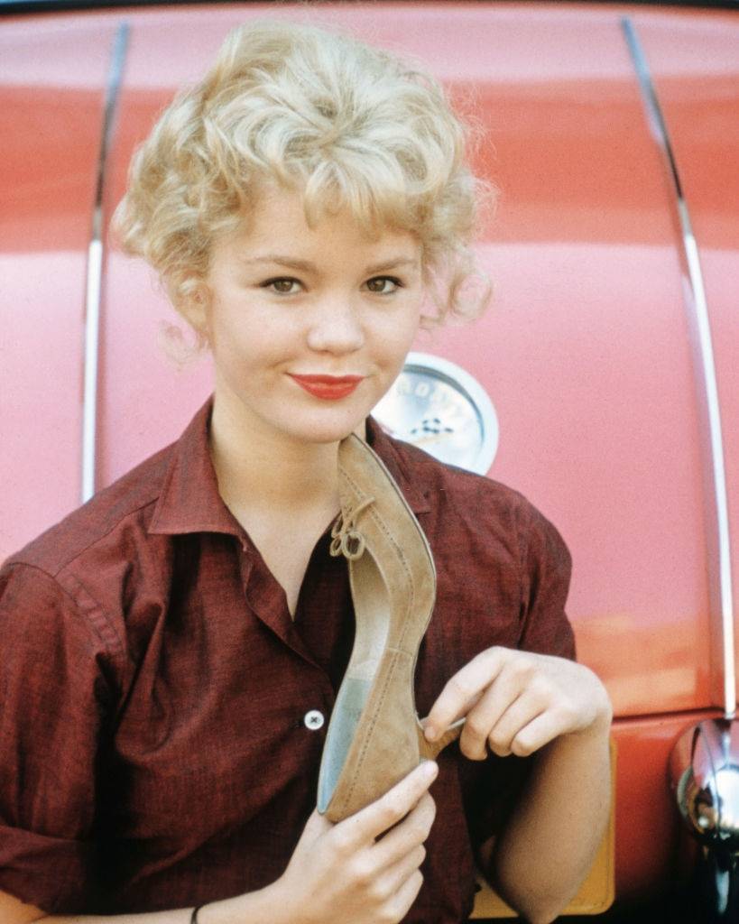 Tuesday Weld wearing a dark red blouse, holding a fawn suede stiletto shoe, 1965.
