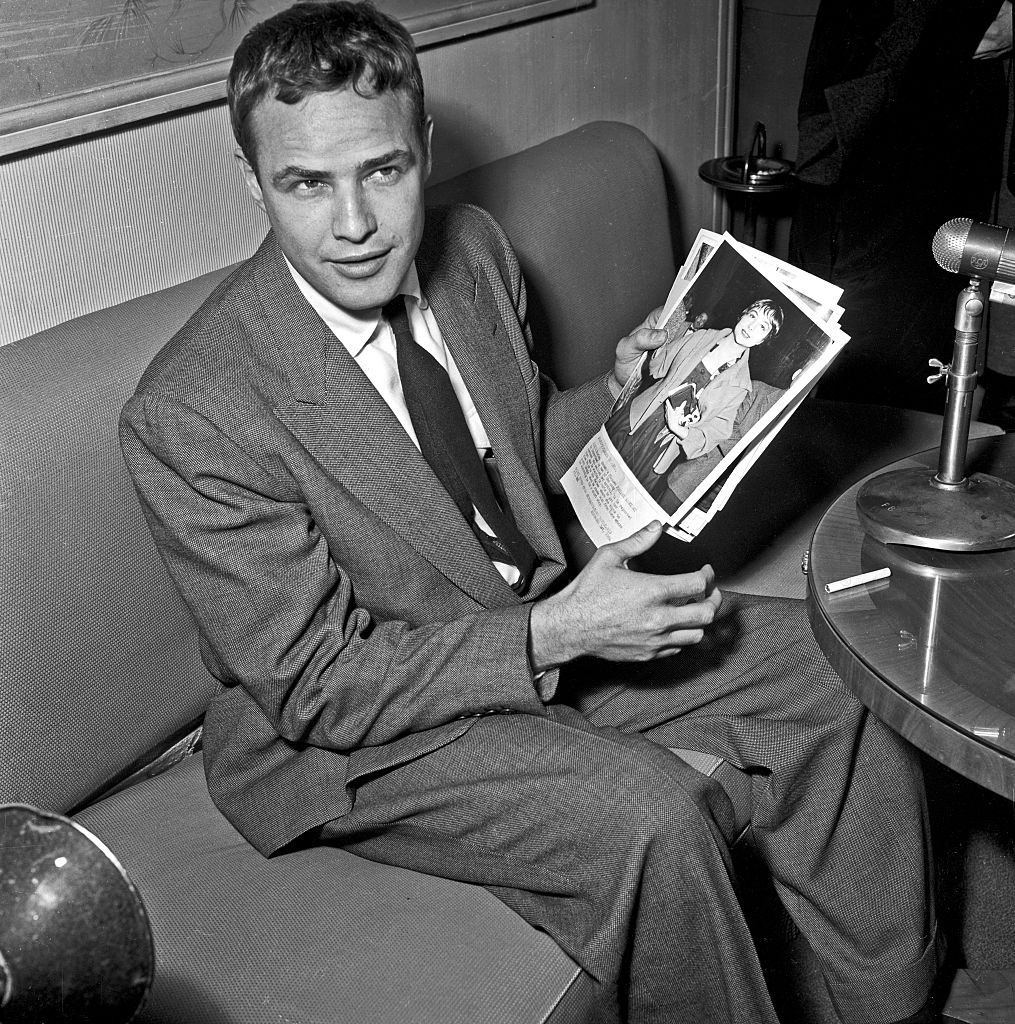 Marlon Brando, sitting on a sofa, showing a photograph during a press conference, 1956.
