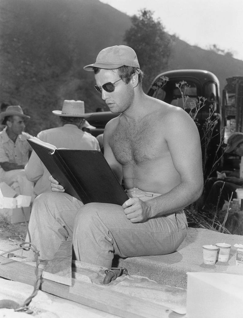 Marlon Brando seated on a block of cement and reading from a book on the outdoor set of the movie, 1953.