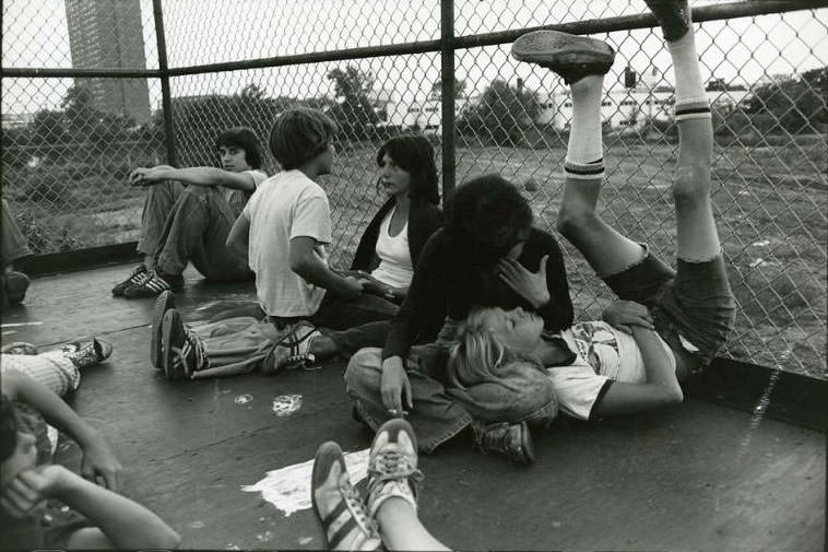 Five teenagers sitting in a fence enclosed area. In the background is the Rindge Towers Housing Project, Jefferson Park, 1973