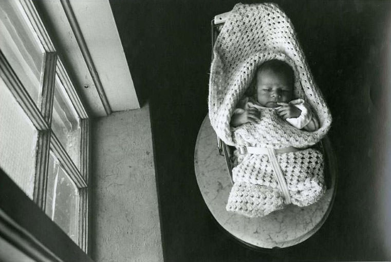 A newborn wrapped in a white blanket, lying in a baby chair, which is on top of a small oval, marble table and net to a window, Jefferson Park, 1975