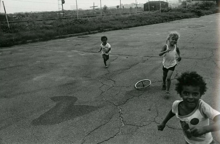 Three children running along a road past a bicycle wheel with spokes lying on the ground. The railroad tracks are visible in the background, Jefferson Park, 1975