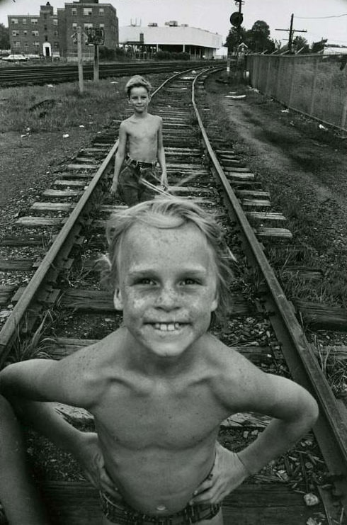 Two boys standing on railroad tracks, both are shirtless. The Jefferson Park Housing Project is visible in the background, Jefferson Park, 1973