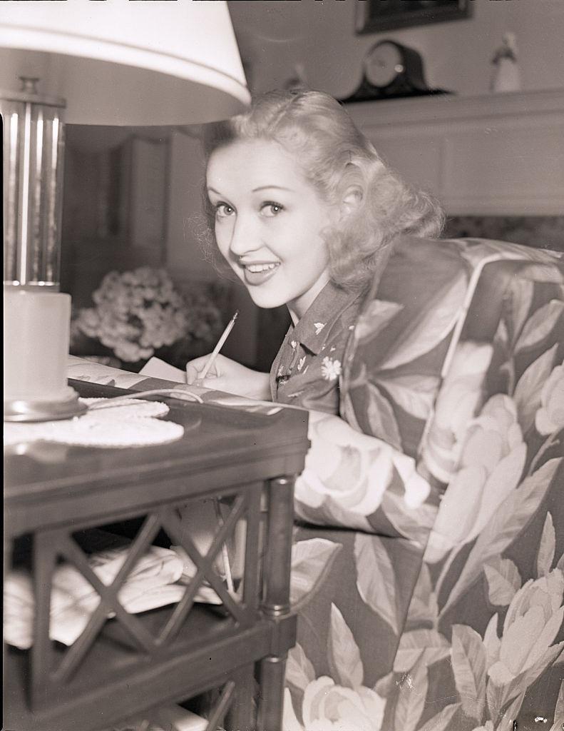 Betty Grable, smiling at the camera while seated and writing in a chair, 1939.