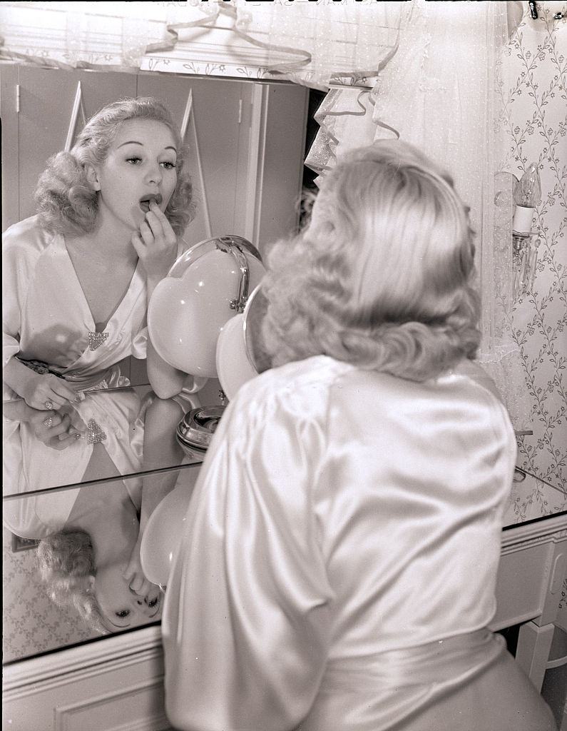 Bertty Grable, wearing a satin robe and applying lipstick in a mirror, 1939.