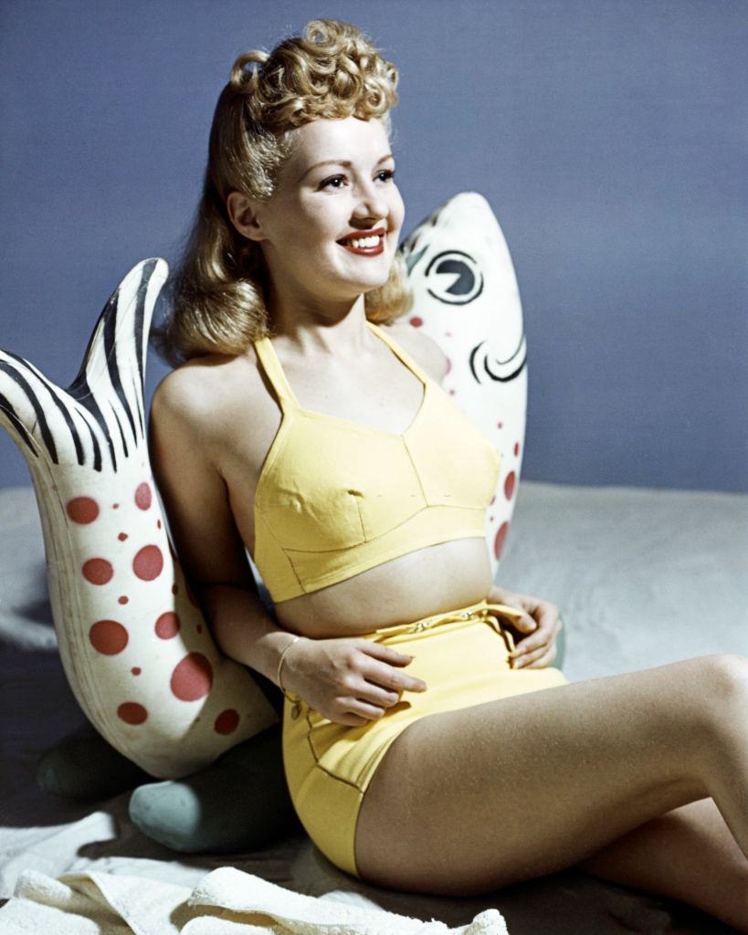 Betty Grable wearing a yellow bikini as she leans against a fish ornament, 1940.