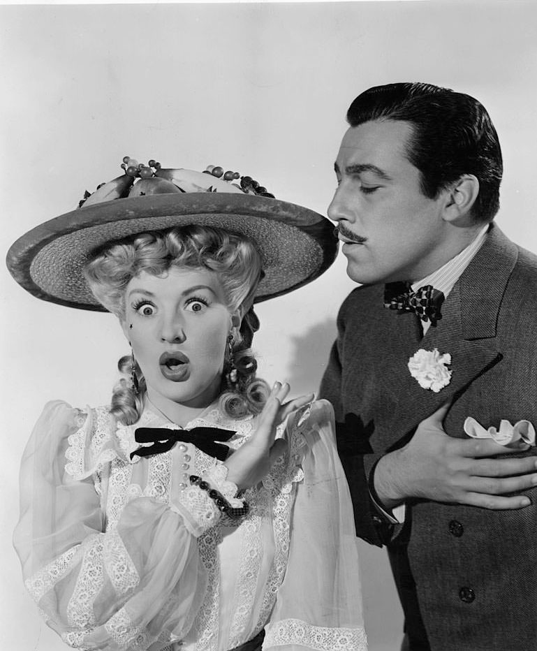 Betty Grable puts her hand up as Cesar Romero holds his heart in a scene from the film 'Coney Island', 1943.