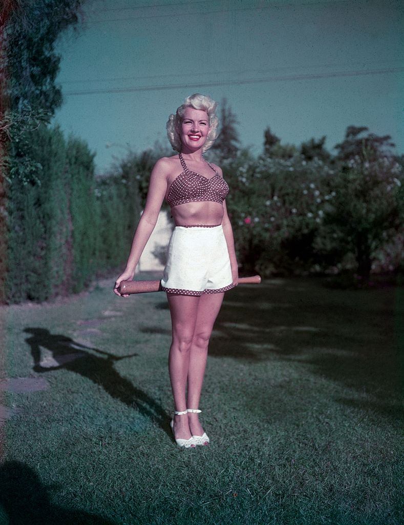 Betty Grable wearing a halter top and shorts while holding a baseball bat on a lawn, 1940s.
