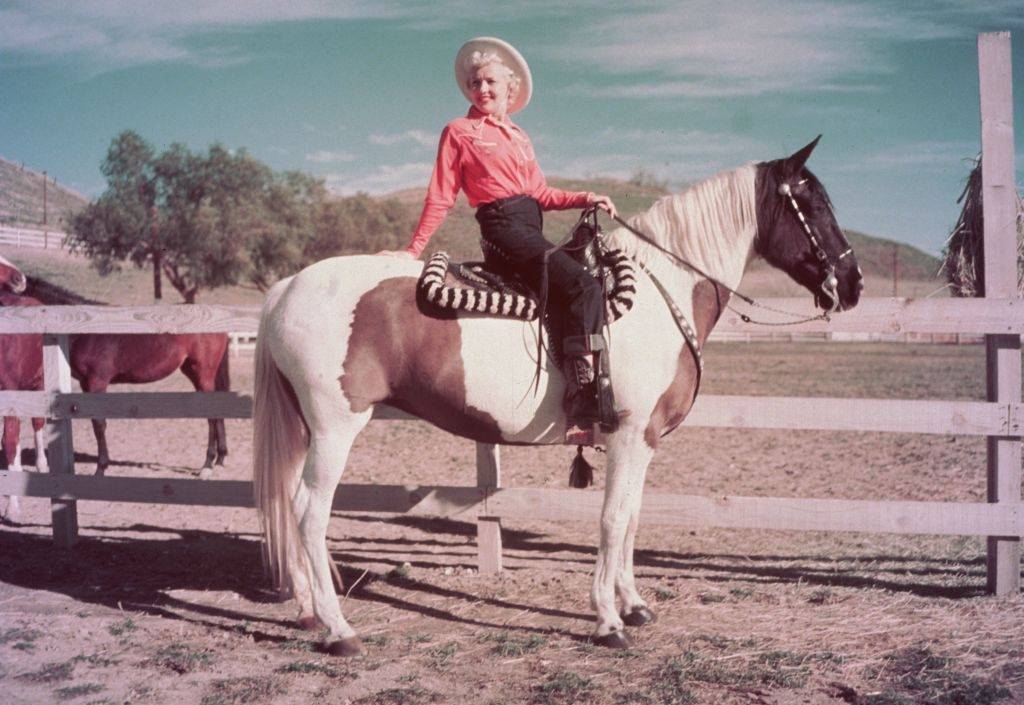 Betty Grable squinting in the sun while seated on a pinto horse with brown patches in a corral, 1952.