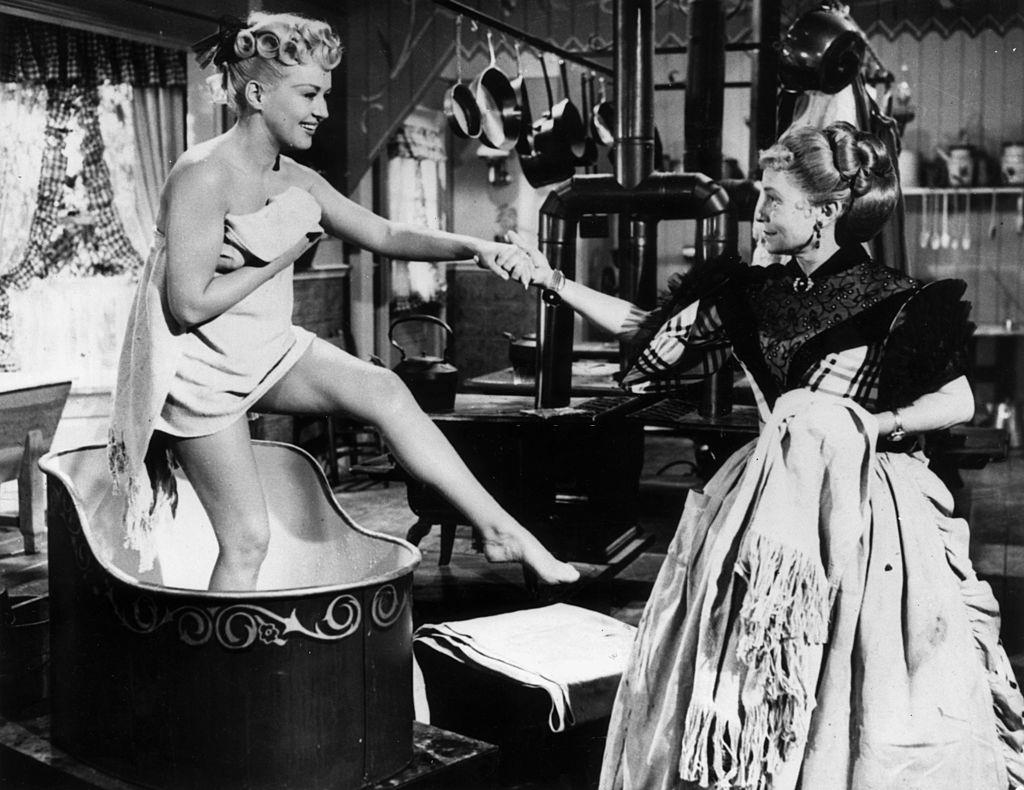 Betty Grable steps out of a wash basin wearing a towel, while Thelma Ritter holds her hand to give her support, 1953.