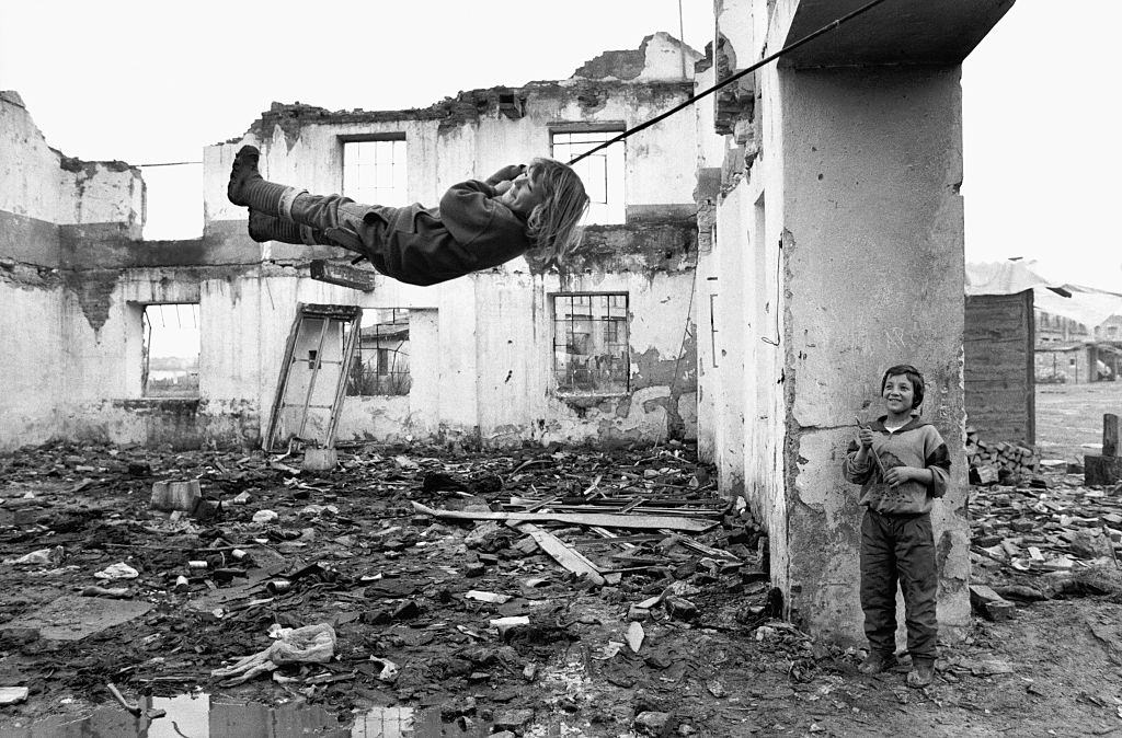 Muslim refugee children swing on a rope swing amongst the rubble in a roofless building.