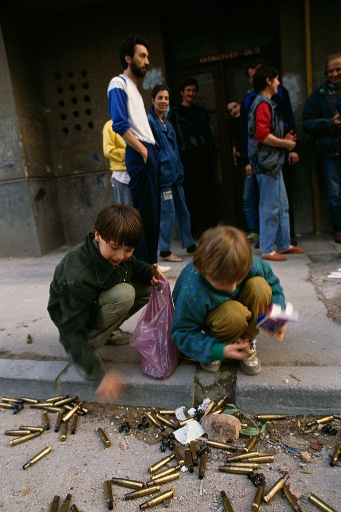 A group of adults watches two Bosnian boys collect machine gun cartridge casings in the streets of Sarajevo while the city is besieged.